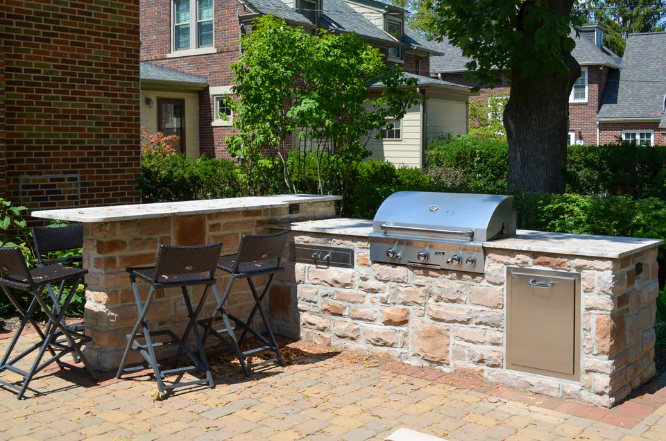 Stone outdoor grill counter with stainless grill, bar seating, brick house, and paver patio